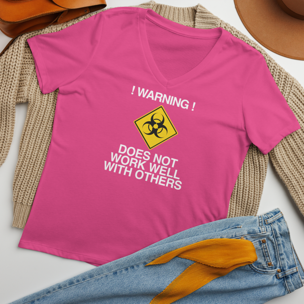 "!Warning! Does Not Work Well With Others" printed on a berry women's relaxed v-neck t-shirt laying on a flat surface amongst other clothes