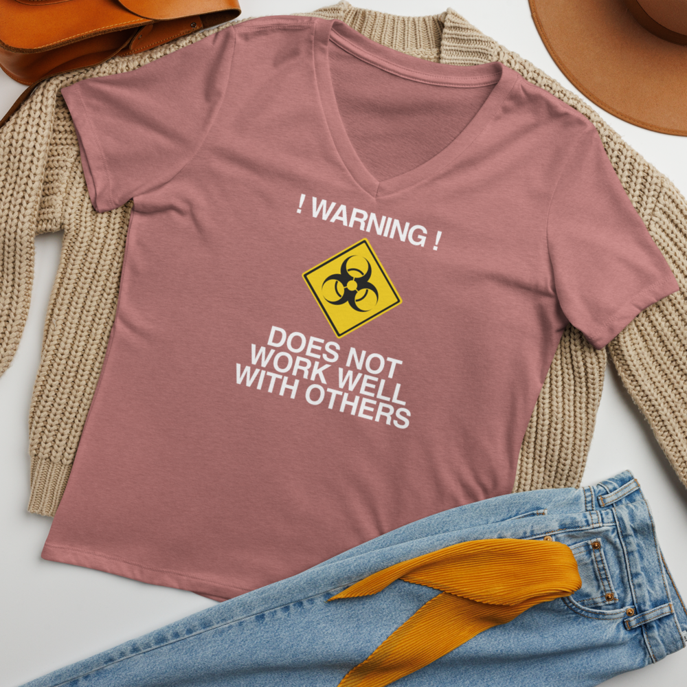 "!Warning! Does Not Work Well With Others" printed on a heather mauve women's relaxed v-neck t-shirt laying on a flat surface amongst other clothes