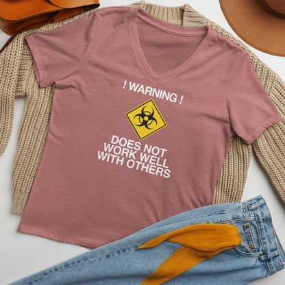 "!Warning! Does Not Work Well With Others" printed on a heather mauve women's relaxed v-neck t-shirt laying on a flat surface amongst other clothes