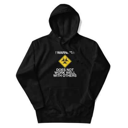 "!Warning! Does Not Work Well With Others" printed on a black unisex hoodie laying on a flat surface with the hood up