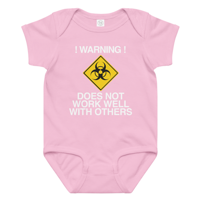 "!Warning! Does Not Work Well With Others" printed on a pink baby jersey bodysuit laying on a flat surface amongst other clothes