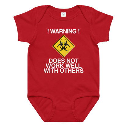 "!Warning! Does Not Work Well With Others" printed on a red baby jersey bodysuit laying on a flat surface amongst other clothes