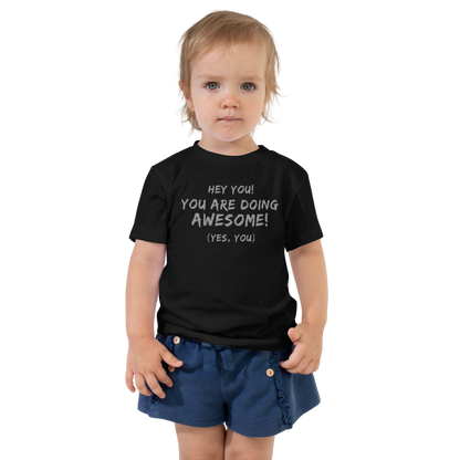 "Hey You! You are doing awesome! (Yes, you)" printed on a black toddler short sleeve tee worn by a female