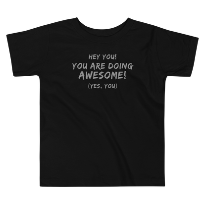 "Hey You! You are doing awesome! (Yes, you)" printed on a black toddler short sleeve tee laying on a flat surface