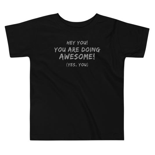 "Hey You! You are doing awesome! (Yes, you)" printed on a black toddler short sleeve tee laying on a flat surface