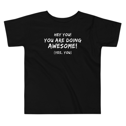 "Hey You! You are doing awesome! (Yes, you)" printed on a black toddler short sleeve tee laying on a flat surface