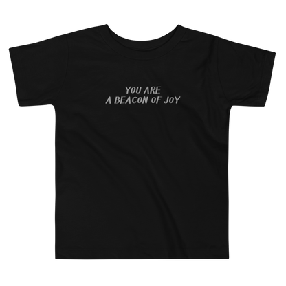 "You are a beacon of joy" printed on a black toddler short sleeve tee laying on a flat surface
