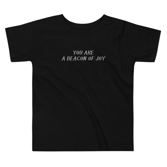 "You are a beacon of joy" printed on a black toddler short sleeve tee laying on a flat surface