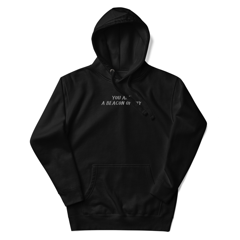 "You are a beacon of joy" printed on a black unisex hoodie laying on a flat surface with the hood up