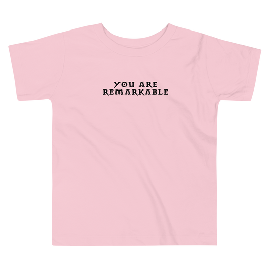 "You are remarkable" printed on a pink toddler short sleeve tee laying on a flat surface