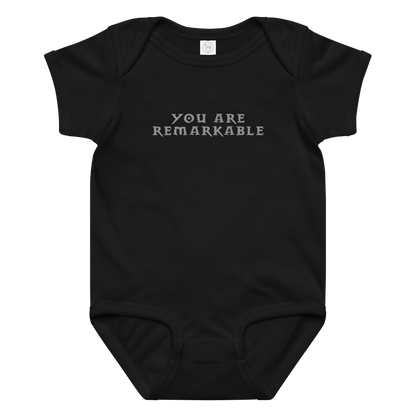 "You are remarkable" printed on a black baby jersey bodysuit laying on a flat surface amongst other clothes