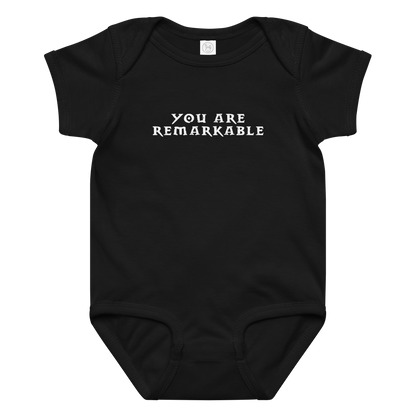 "You are remarkable" printed on a black baby jersey bodysuit laying on a flat surface amongst other clothes