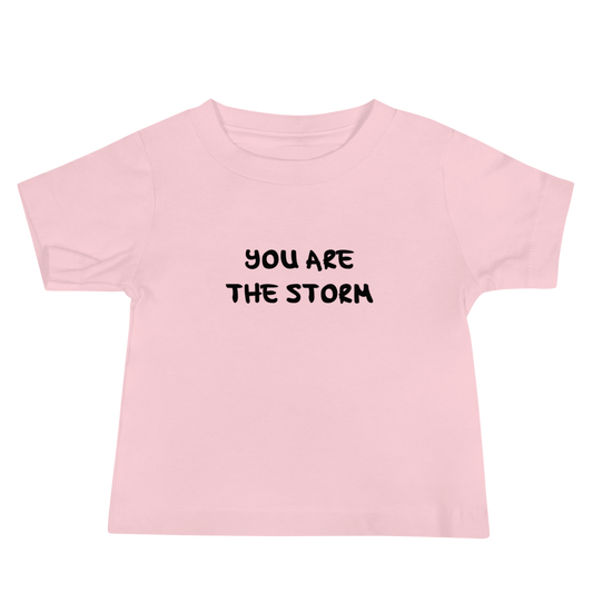"You are the storm" printed on a pink baby jersey short sleeve tee laying on a flat surface