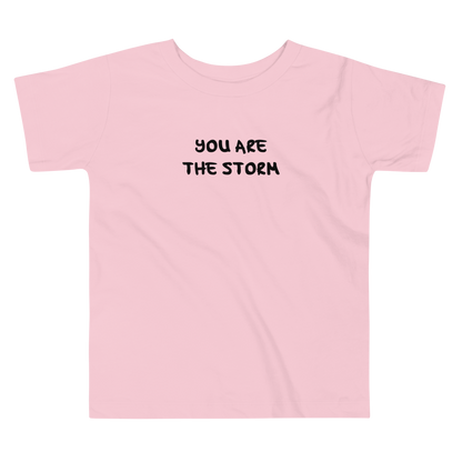 "You are the storm" printed on a pink toddler short sleeve tee laying on a flat surface