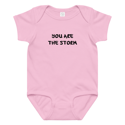 "You are the storm" printed on a pink baby jersey bodysuit laying on a flat surface amongst other clothes