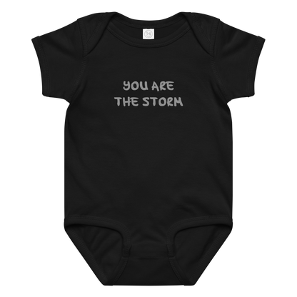 "You are the storm" printed on a black baby jersey bodysuit laying on a flat surface amongst other clothes