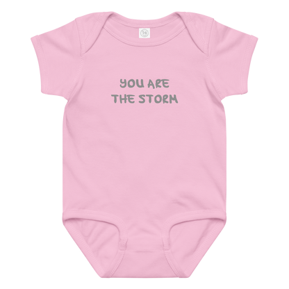 "You are the storm" printed on a pink baby jersey bodysuit laying on a flat surface amongst other clothes