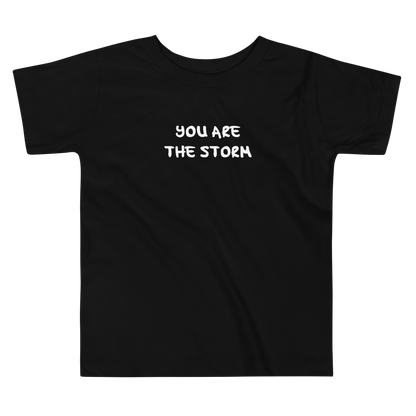 "You are the storm" printed on a black toddler short sleeve tee laying on a flat surface