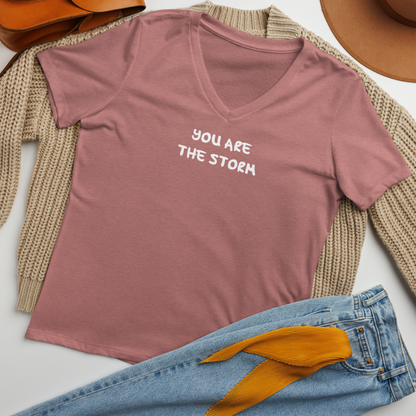 "You are the storm" printed on a heather mauve women's relaxed v-neck t-shirt laying on a flat surface amongst other clothes